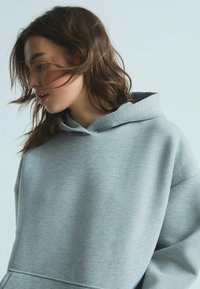 Grå, overdimensioneret hoodie lavet af blødt stof med hætte. Har en kengurulomme og ribbede manchetter, hvilket giver en tekstureret finish.