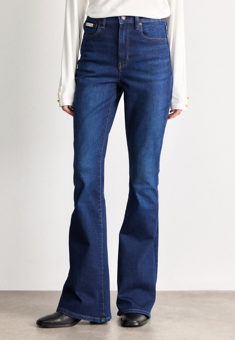 Τζιν με ψηλή μέση και καμπάνα σε σκούρο μπλε τζιν ύφασμα, με μπροστινές τσέπες, διακριτική ραφή και απαλή υφή. Συνδυασμένο με μαύρα παπούτσια.