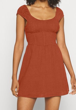 Vrouw met een kledingsstuk met korte mouwen, roestkleurige, nauwsluitende taillejurk met een vierkante halslijn, staand met armen ontspannen naast het lichaam tegen een lichte achtergrond.