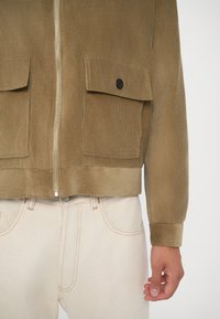 Brūnas korduroja jakas ar rāvējslēdzēju, divām priekšējām kabatām, ribota tekstūra un kontrastējoša melna poga, kombinēta ar gaišiem džinsiem.