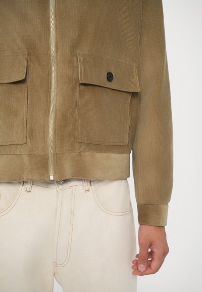 Giacca in corduroy marrone con chiusura a zip, due tasche frontali, texture a coste e bottone nero a contrasto, abbinata a jeans chiari.