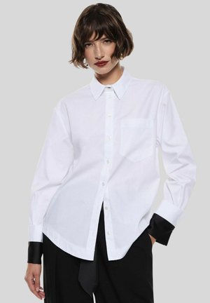 Camicia bianca con colletto classico, maniche lunghe con polsini neri e taschino frontale, realizzata in tessuto liscio.