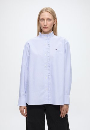Kvinde med lyst, blond, glat hår, iført en lyseblå, striped skjorte med knapper og flæsekrave samt sorte bukser mod en ensartet hvid baggrund.