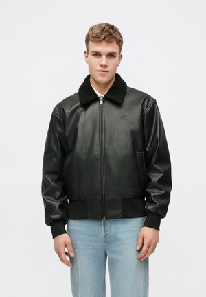 Νεαρός άνδρας με μαύρο δερμάτινο μπουφάν και κολάρο από φλις, φορώντας αμάνικα μπλε τζιν, που στέκεται μπροστά σε λευκό φόντο.