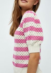 Žena v růžovém a bílém pruhovaném háčkovaném topu s nadýchanými krátkými rukávy, ukázaná z boku na jednobarevném pozadí.