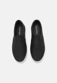 Sorte slip-on sko med teksturert mønster, gummisåle og minimalistisk design. Har logo på innersålen og glatt finish.