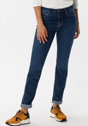 Jeans de mezclilla azul oscuro con corte recto, cinco bolsillos, costuras sutiles y puños enrollados. Combinados con zapatillas deportivas naranjas y negras.