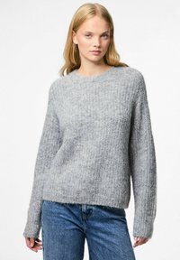 Jauna sieviete ar garām blondām matēm, tērpusies ar teksturētu gaiši pelēku dziju džemperi un ziliem džinsa biksēm, stāv pretī baltai fonam.