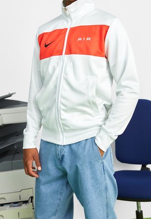 Personne portant une veste blanche zippée avec un panneau orange sur la poitrine et un logo Swoosh noir, debout près d'une imprimante de bureau et d'une chaise bleue.