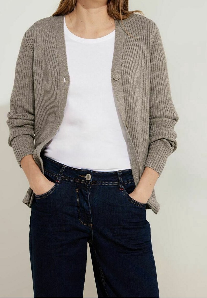 Personne portant un cardigan côtelé beige par-dessus une chemise blanche, les mains dans les poches d'un jean bleu foncé.