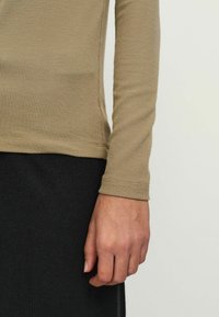 Hosszú ujjú felső bordázott bézs anyagból, testhezálló szabással. A közelkép egy kéz pihenését mutatja egy fekete textúrájú szoknyán.