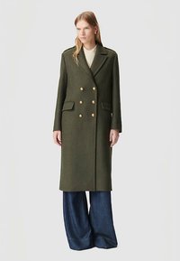 Kobieta ubrana w długi oliwkowozielony płaszcz dwurzędowy z złotymi guzikami i szerokie, ciemnoniebieskie spodnie, stojąca na jednolitym tle.