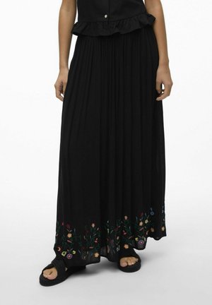 Schwarzer Maxikleid aus fließendem Stoff mit buntem Blumenstickerei am Saum; kombiniert mit strukturierten schwarzen Sandalen.