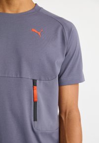 Gros plan d'une personne portant un t-shirt gris Puma avec un logo orange et une fermeture éclair noire verticale avec un tire-zip orange.