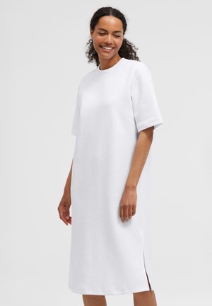 Lächelnde Frau mit lockigem Haar, die ein schlichtes weißes Midikleid mit kurzen Ärmeln und seitlichen Schlitzen trägt, vor hellem Hintergrund stehend.