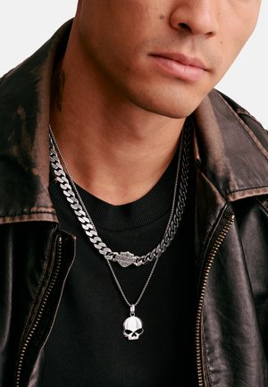 Nahaufnahme einer Person, die eine schwarze Lederjacke, ein schwarzes Hemd und zwei silberne Halsketten trägt, eine mit einem Totenkopf-Anhänger und eine mit dem Harley-Davidson-Logo.