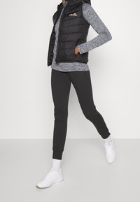 Svart pufferväst över en grå långärmad topp, matchad med svarta tapered joggers och vita sneakers. Västet har en logotyp på bröstet.