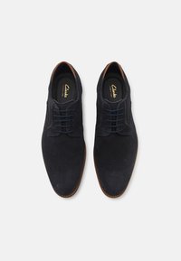 Scarpe da uomo in suede blu navy con una texture liscia, punta rotonda e dettagli in pelle marrone. Presentano lacci tradizionali e una soletta imbottita.