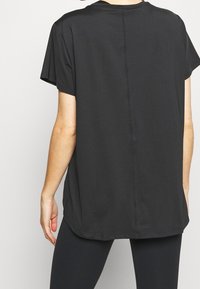 Persona con una camiseta negra de manga corta y holgada y leggins negros, vista desde atrás contra un fondo claro y sencillo.