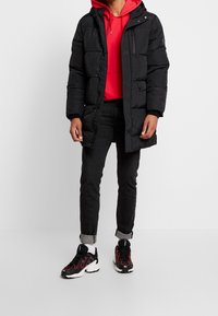 Černá péřová bunda s kapucí, předním zipem a bočními kapsami, nošená přes červenou mikinu s kapucí a černé džíny, doplněná černo-červenými teniskami.