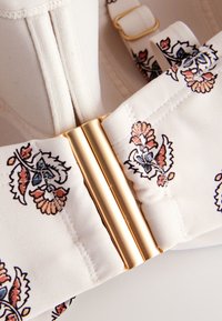 Fermoir doré sur tissu crème avec motif floral brodé en bleu, marron et orange, attaché à un vêtement blanc texturé.