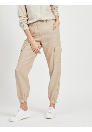 Persoon draagt beige cargobroek met elastische boorden en witte sneakers, staat met één hand in de zak en de andere op de dij.