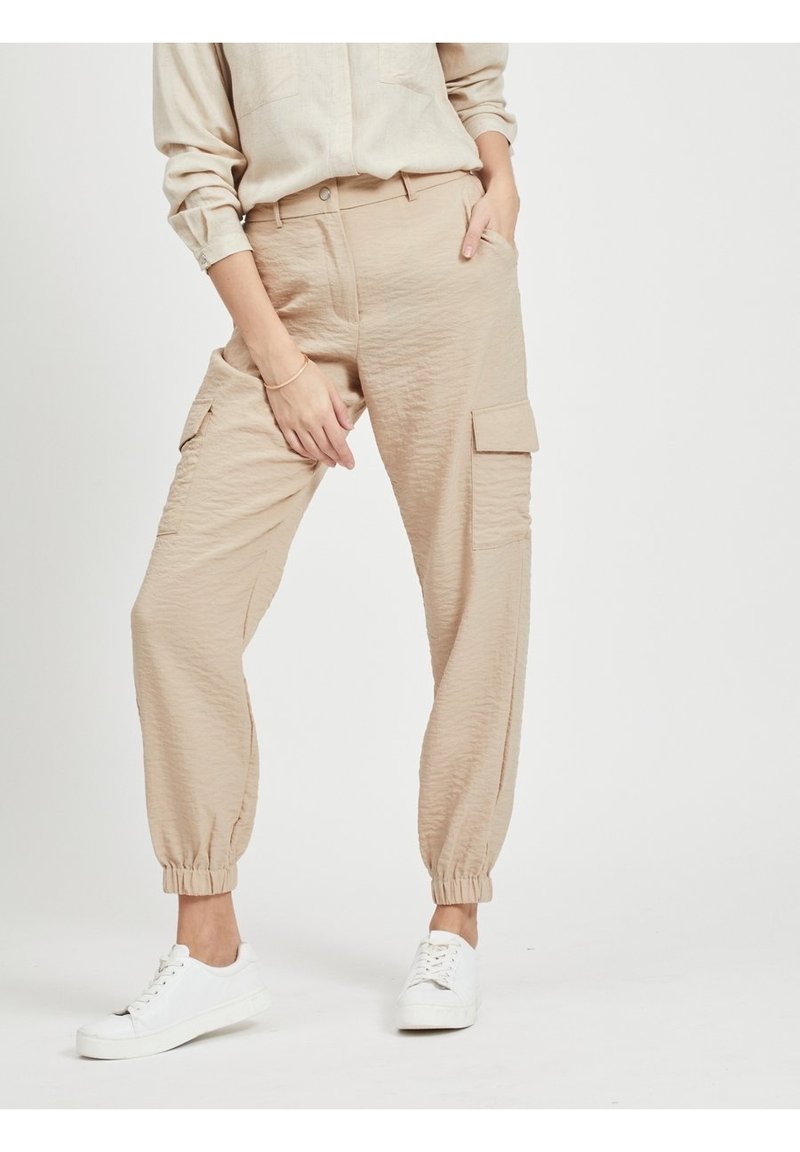 Personne portant un pantalon cargo beige avec des bas élastiques et des baskets blanches, debout avec une main dans la poche et l’autre sur la cuisse.
