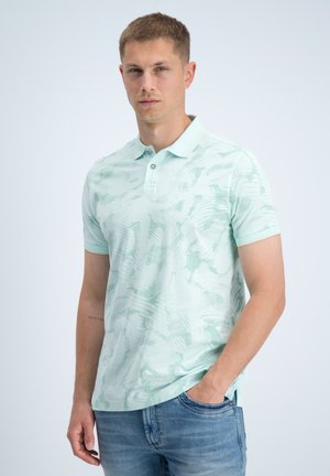Uomo con polo verde chiaro a fantasia e jeans blu, in piedi con una mano in tasca su sfondo uniforme.