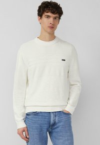 Jeune homme aux cheveux bouclés portant un pull blanc texturé et un jean bleu, debout avec une main dans la poche devant un fond uni.