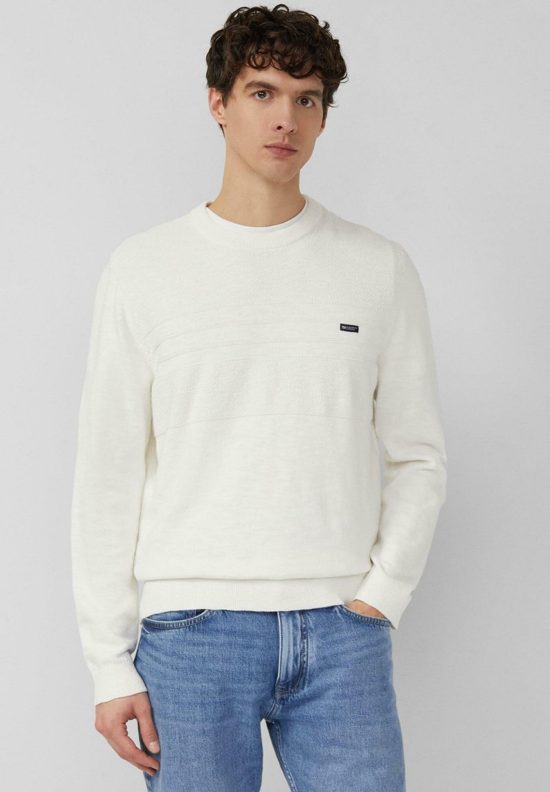 Jeune homme aux cheveux bouclés portant un pull blanc texturé et un jean bleu, debout avec une main dans la poche devant un fond uni.