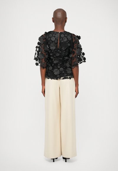 Sort top med blomstermotiv broderet, der har gennemsigtige ærmer, sammen med cremede vide bukser. Toppen har et nøglehul i ryggen og strukturerede stofdetaljer.