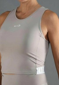 Primer plano de una persona que lleva un top corto sin mangas de color gris claro con un pequeño símbolo de infinito blanco en el pecho y una cinturilla elástica blanca.