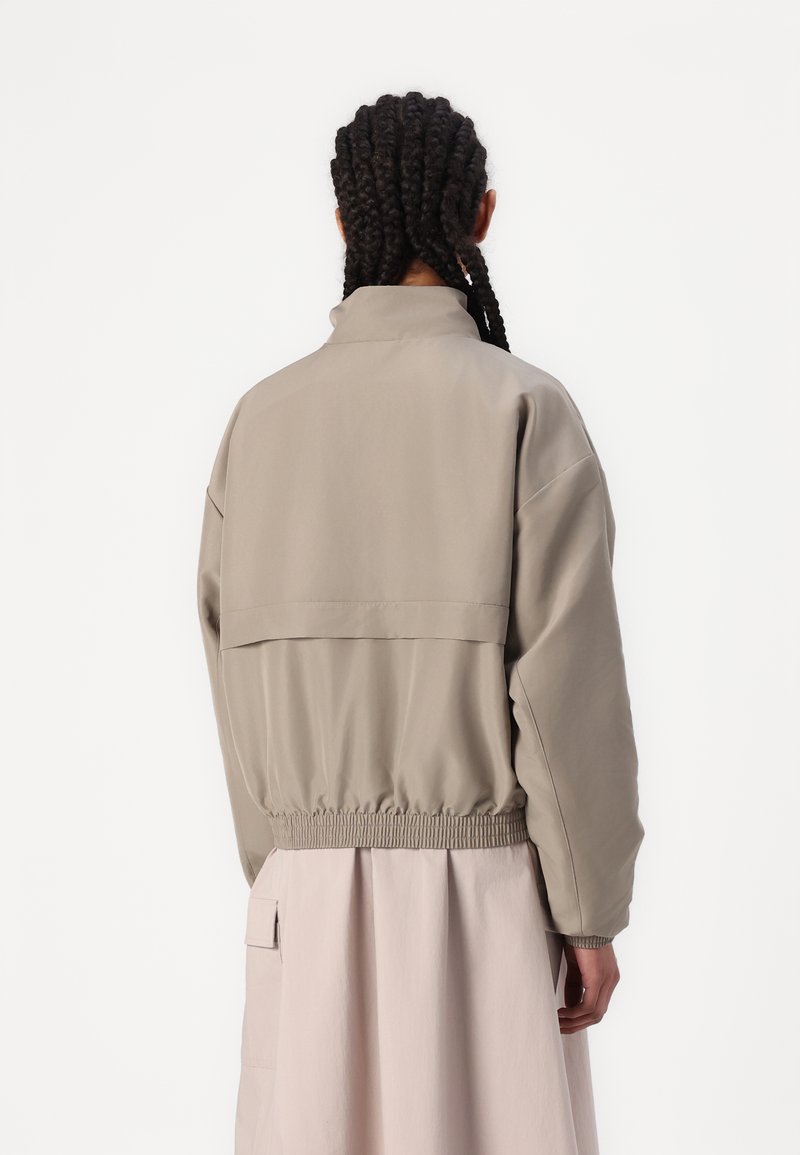 Veste légère beige avec un col montant, une taille élastique et des épaules tombantes ; présente un design de yoke au dos. Portée par-dessus une jupe rose.