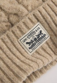 Beige gerippte Strickmütze mit einem texturierten Design, versehen mit einem gewebten Etikett, auf dem "Levi Strauss & Co." und Pferdegrafiken abgebildet sind.