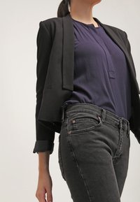 Blazer noir sur mesure porté sur un chemisier bleu marine, associé à un jean en denim gris taille haute, slim, avec des coutures discrètes et des accents métalliques.