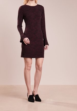 Kobieta ubrana w dopasowaną, ciemnoczerwoną sukienkę na długi rękaw z subtelnym wzorem oraz czarne mules na obcasie, stojąca na jednolitej podłodze.