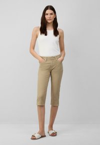 Pantaloni beige a vita alta con una vestibilità slim, abbinati a una canotta bianca a costine. Calzature sono sandali bianchi slip-on con un accento decorativo.