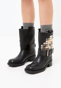 Botines de cuero negro con acabado texturizado, detalles de tachuelas plateadas en el lado exterior, banda ancha en el tobillo y tacón bajo cuadrado. Medias grises visibles.