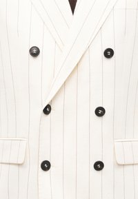 Λευκό μπλέιζερ διπλής σειράς με λεπτές καφέ κάθετες ρίγες, μαύρα κουμπιά, γιακά με άνοιγμα και τσέπες με πατιλέτες στο ύψος της μέσης.