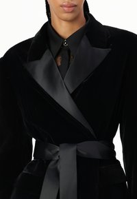 Czarna aksamitna marynarka z satynowym kołnierzem szalowym, pasującym satynowym paskiem i koronkowym detalem pod kołnierzem. Teksturowany materiał o dopasowanym kroju.