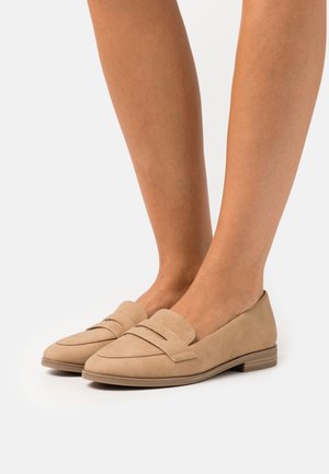 Pár béžových nízkých semišových mokasín nošených na holou nohu, s páskovým detailem přes horní část vpředu, na bílém pozadí.
