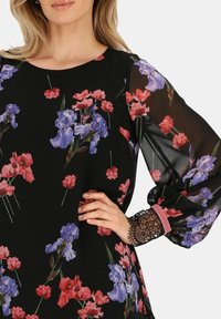 Kwiatowa bluzka na czarnym tle, ozdobiona fioletowymi i czerwonymi wzorami kwiatów. Przezroczyste rękawy z koronkowymi akcentami i różowym wykończeniem przy mankietach.