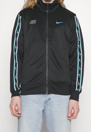 Chaqueta de chándal negra de Nike con cuello alto, cremallera frontal y franjas laterales turquesas con patrones de logotipos repetidos. Textura de tela suave.
