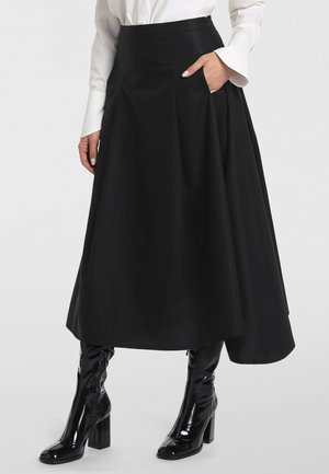 Femme portant une jupe midi noire taille haute, évasée en A, avec la main dans la poche, un chemisier blanc et des bottes à talons en cuir verni noir.