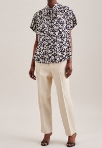 Blommig blus i marinblått med krämfärgade blommor, korta ärmar och rosettkrage. Matcher med krämfärgade, texturerade vida byxor och svarta klackar.