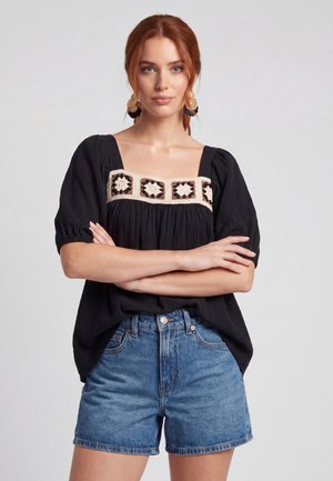 Femme aux cheveux roux portant un chemisier noir avec une broderie florale et un short en jean bleu, debout les bras croisés devant un fond uni.