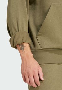 Olivovo zelená mikina s nariadenými rukávmi a prednou vreckom; mäkká tkanina s hladkou textúrou. Viditeľné tetovanie na zápästí.