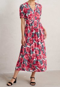 Kvinna som bär en rosagrå blommönstrad V-ringad midiklänning med korta ärmar och svarta sandaletter med ankelsnörning, stående inomhus.