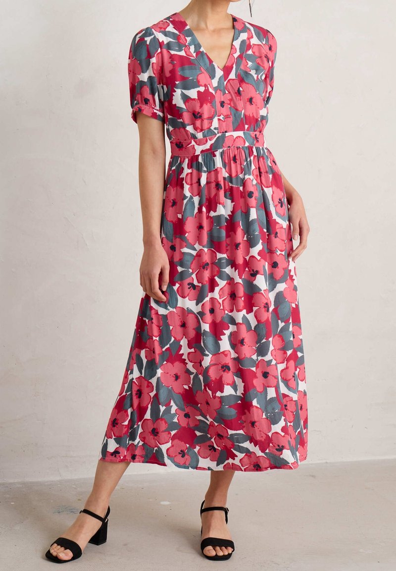 Kvinna som bär en rosagrå blommönstrad V-ringad midiklänning med korta ärmar och svarta sandaletter med ankelsnörning, stående inomhus.