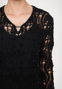 Top in pizzo nero con intricati motivi a foglia, maniche lunghe, scollatura a V e dettagli perlacei per un effetto tattili aggiuntivo.